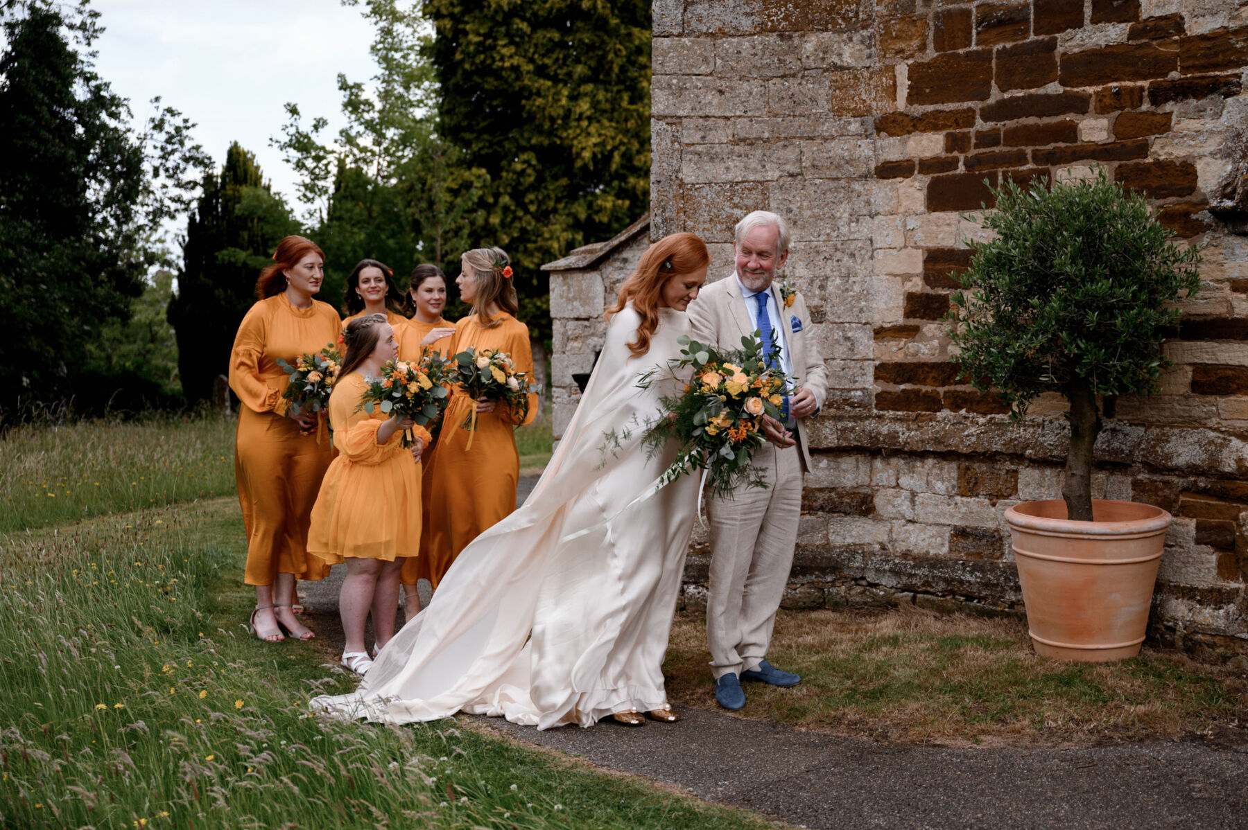 Bride in Cortana wedding dress waiting to enter the church with her father. Bridesmaids in orange dresses. Taylor Hughes Photography.