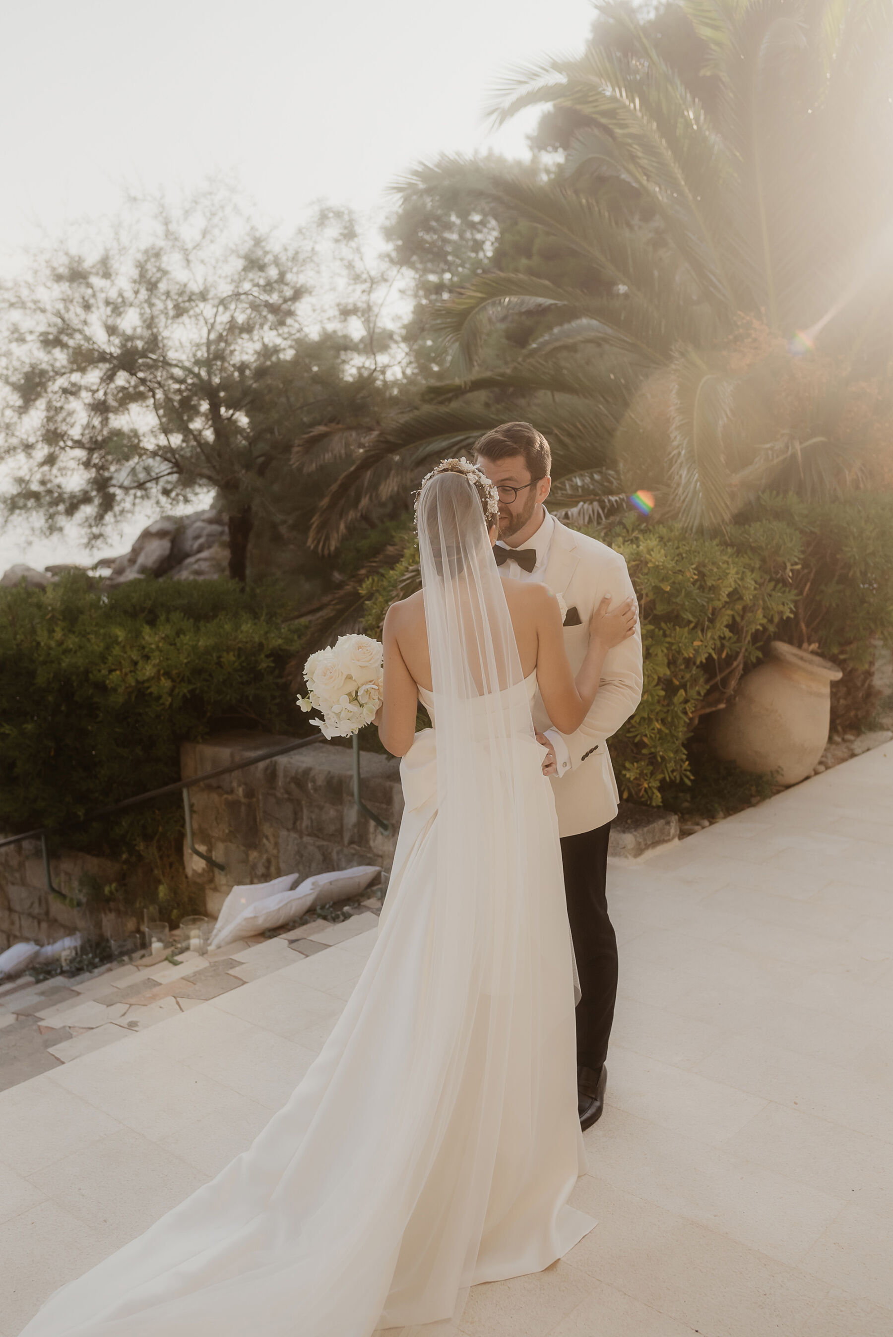 Sarah in Eva Lendel for her Classy & Contemporary Wedding in Croatia 23 Groom in white tux kissing bride in minimalist wedding dress and long veil carrying a bouquet of all white flowers