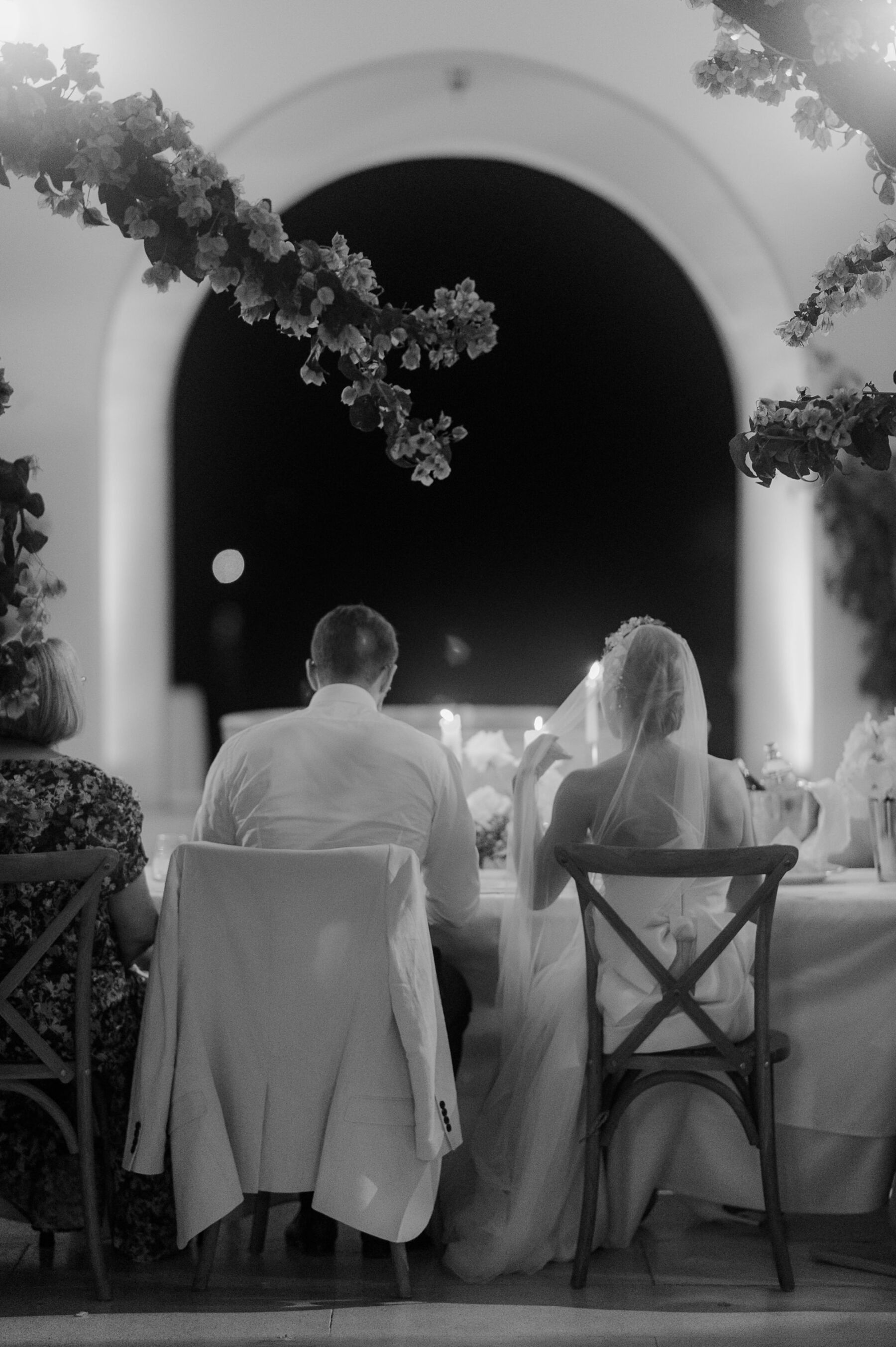Sarah in Eva Lendel for her Classy & Contemporary Wedding in Croatia 45 Shot from behind of bride and groom sitting next to one another on cross back wooden chairs with an archway head of them at night time.