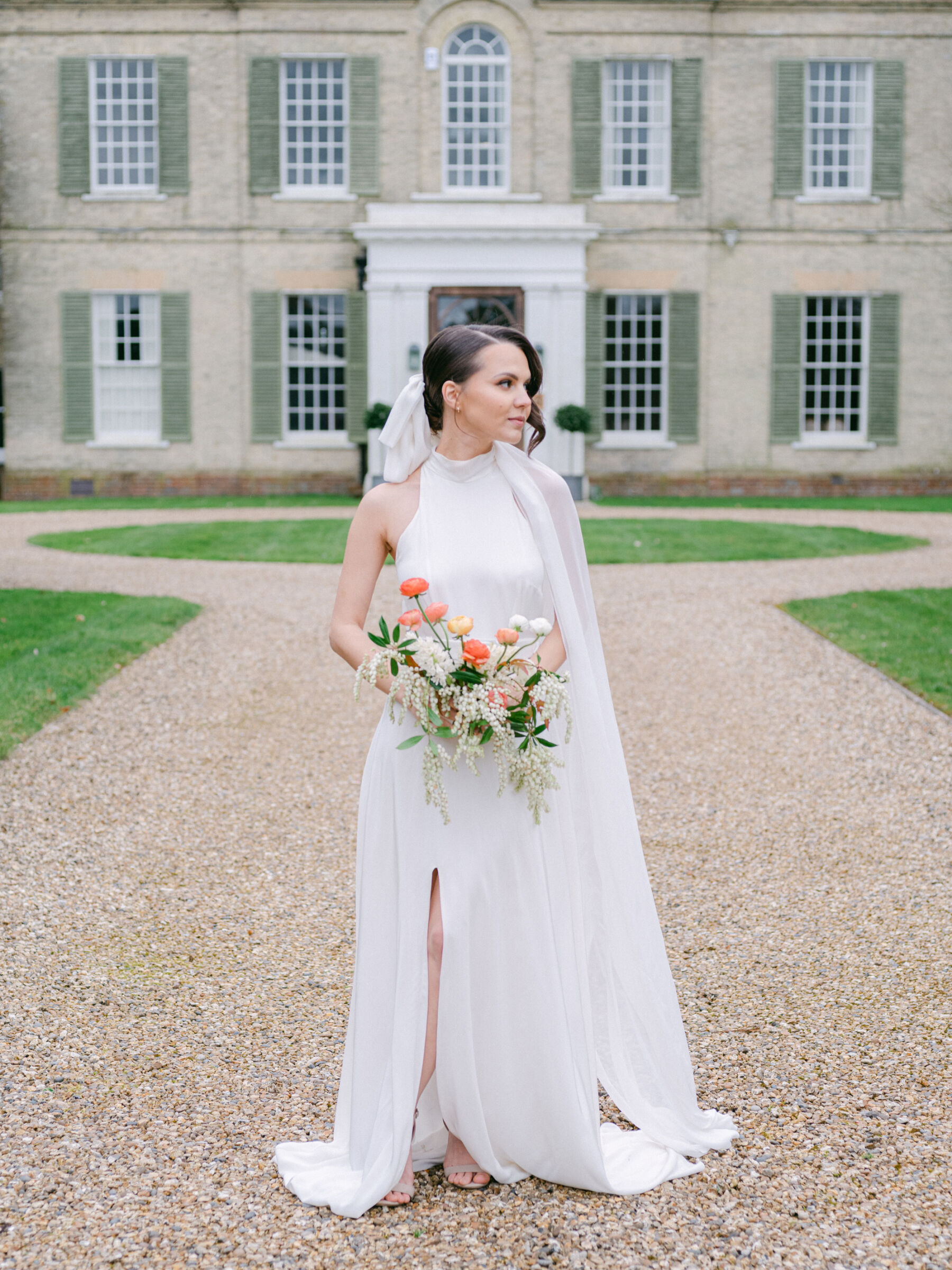 Celebrating Seasonal, British Grown Wedding Flowers With Flower & Fern at Findon Place 17 Bride standing in the grounds of Findon Place wedding venue wearing a halterneck wedding dress with a thigh length slit. The bride carries a bouquet of British grown Ranunculus and seasonal flowers.