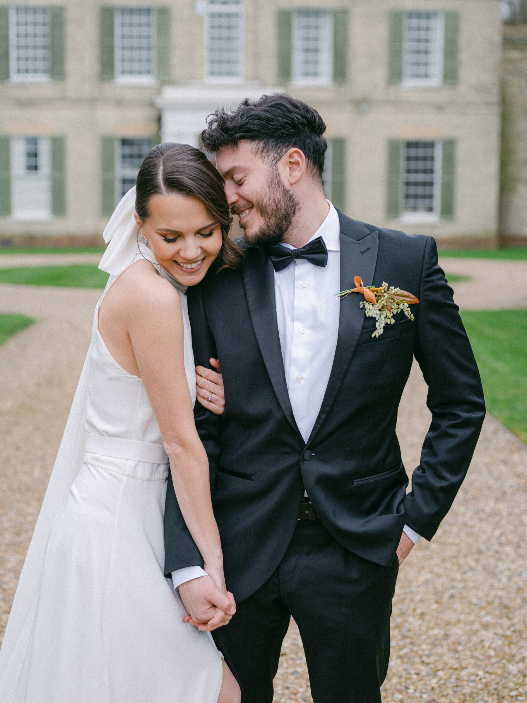 Celebrating Seasonal, British Grown Wedding Flowers With Flower & Fern at Findon Place 22 Groom in a bow tie and tuxedo smiling and nuzzling affectionally into the hair of his bride wearing a halterneck wedding dress.