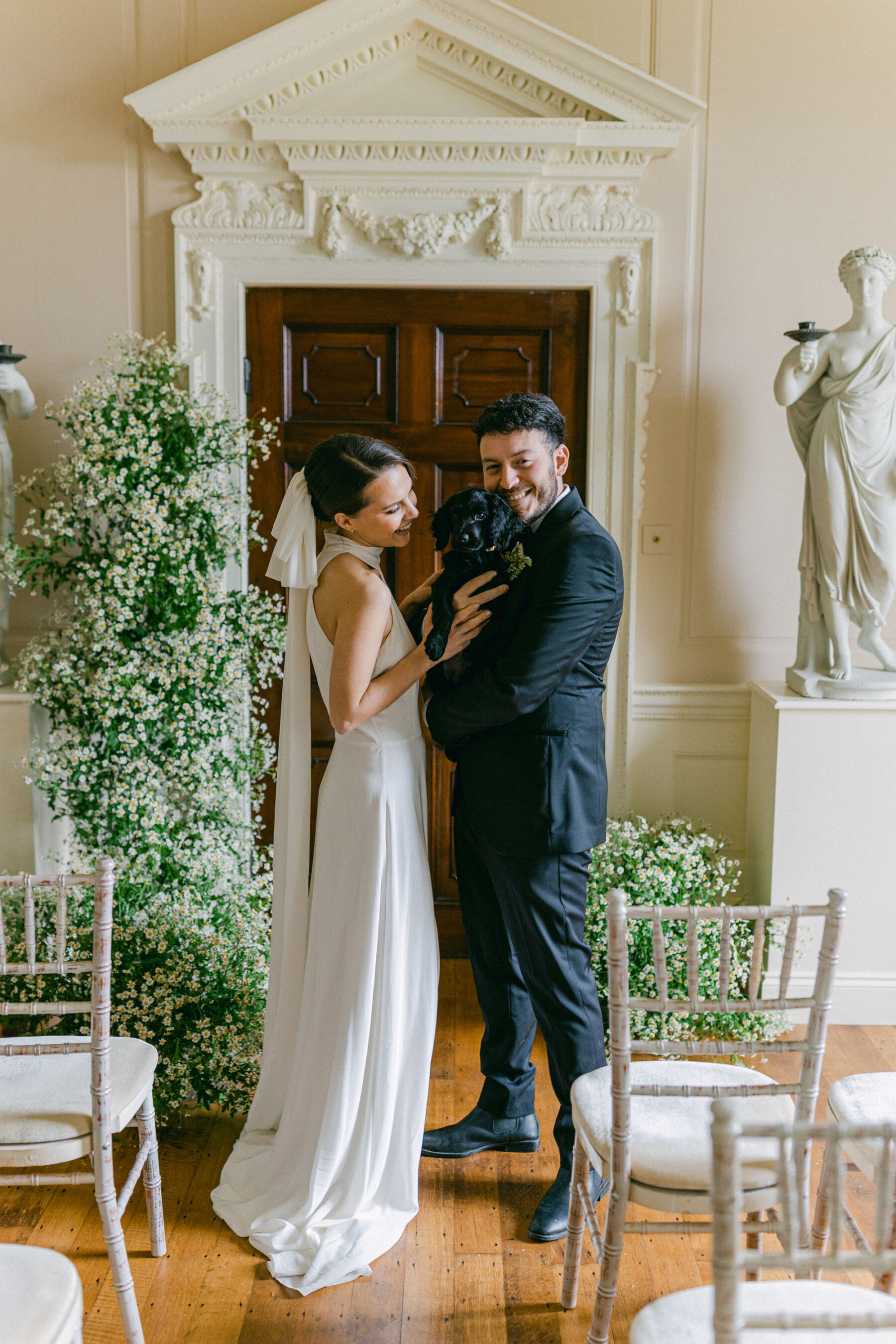 Celebrating Seasonal, British Grown Wedding Flowers With Flower & Fern at Findon Place 24 Bride and groom standing inside Findon Place next to a beautiful daisy floral arch holding a cute little black dog.
