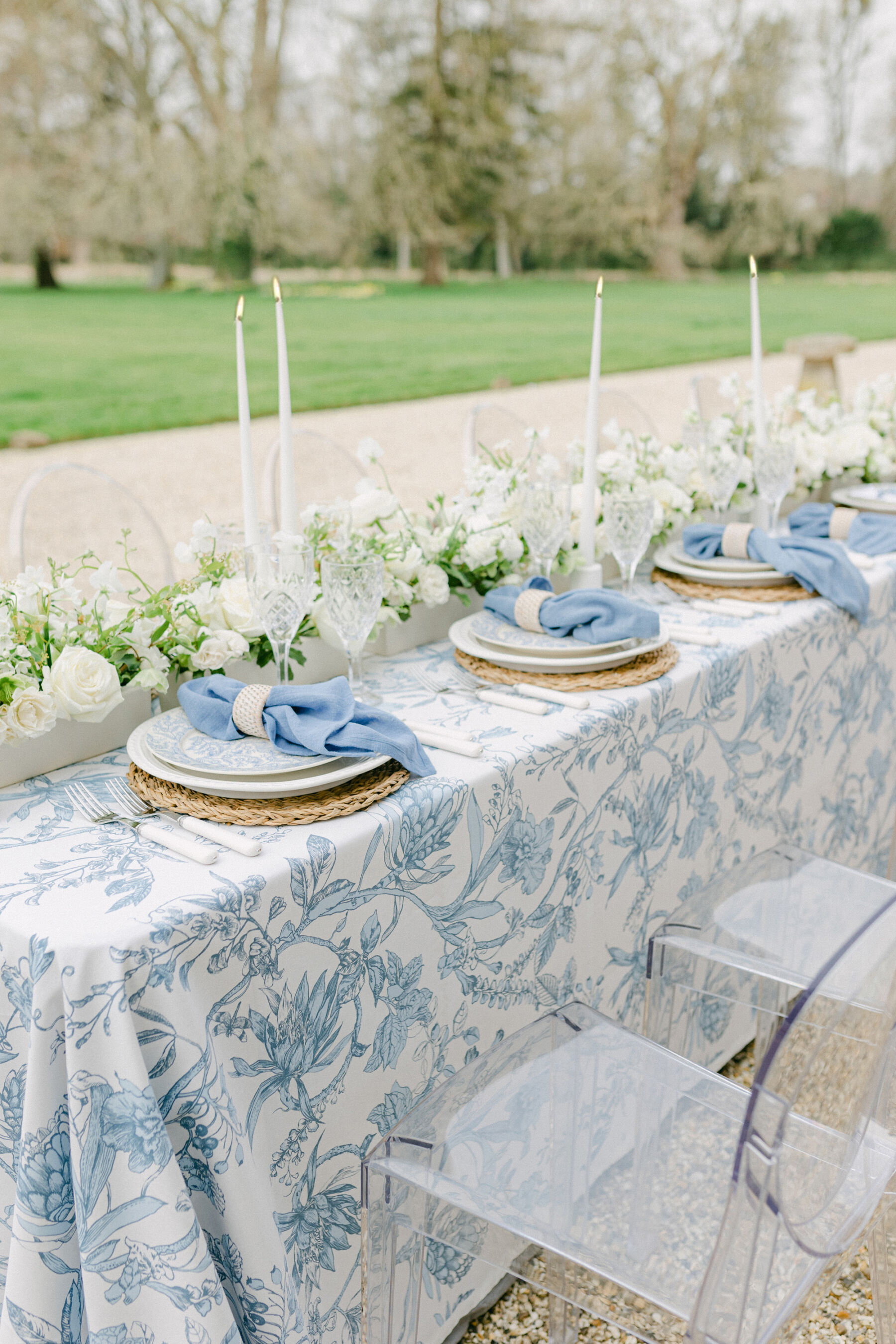 Pale Baby Blue Spring Wedding Inspiration at Avington Park, Featuring Halfpenny London 3 Outdoor wedding reception table setting with patterned blue table cloth, pale blue napkins and tall taper candles. A long and low running of pale white flowers provides floral decor down the centre of the table which is lined with Ghost chairs.