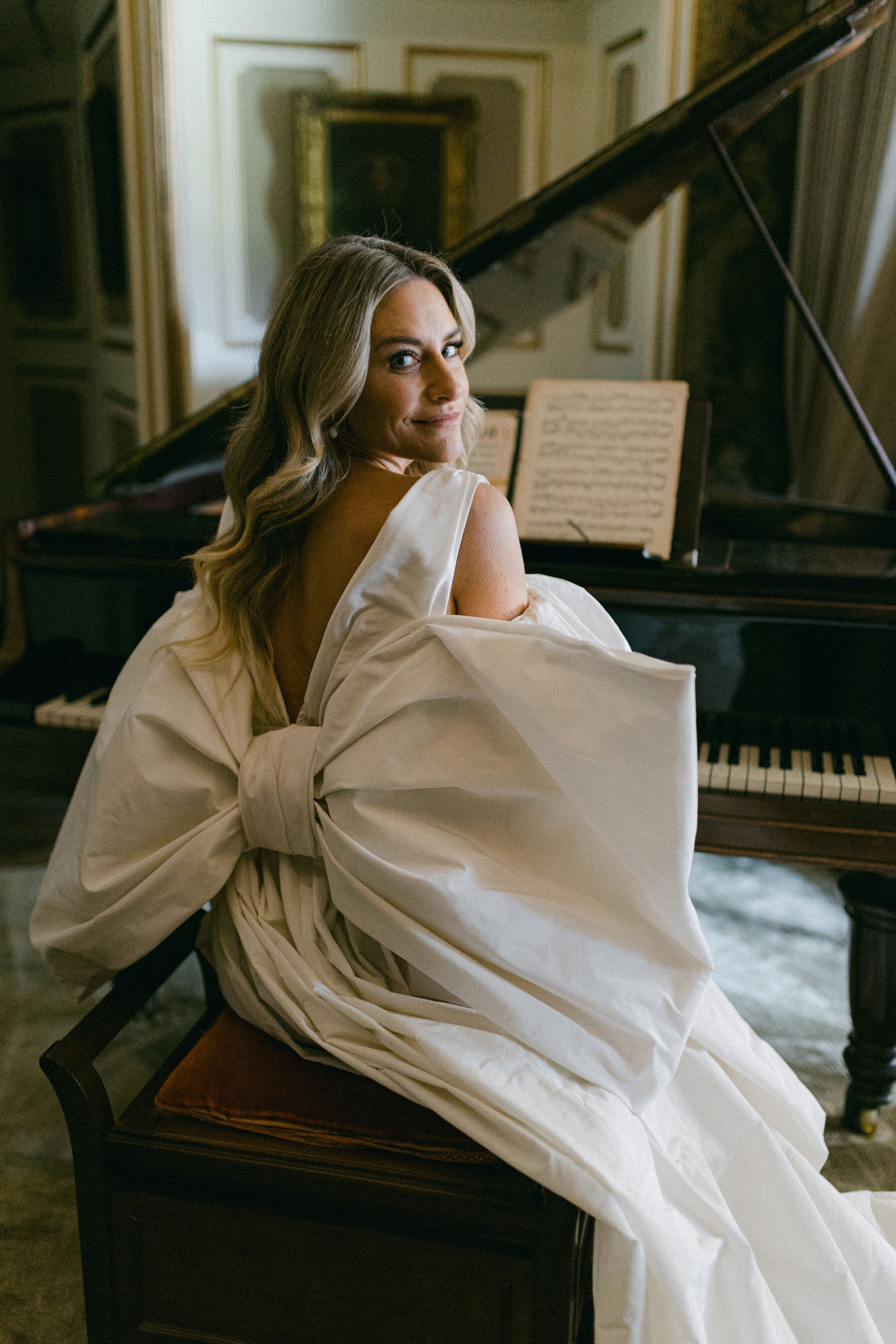 Pale Baby Blue Spring Wedding Inspiration at Avington Park, Featuring Halfpenny London 18 Big bow.Wedding dress with a huge statement bow at the back, by Halfpenny London. Bride is sat at the piano and turning around to to look at the viewer.