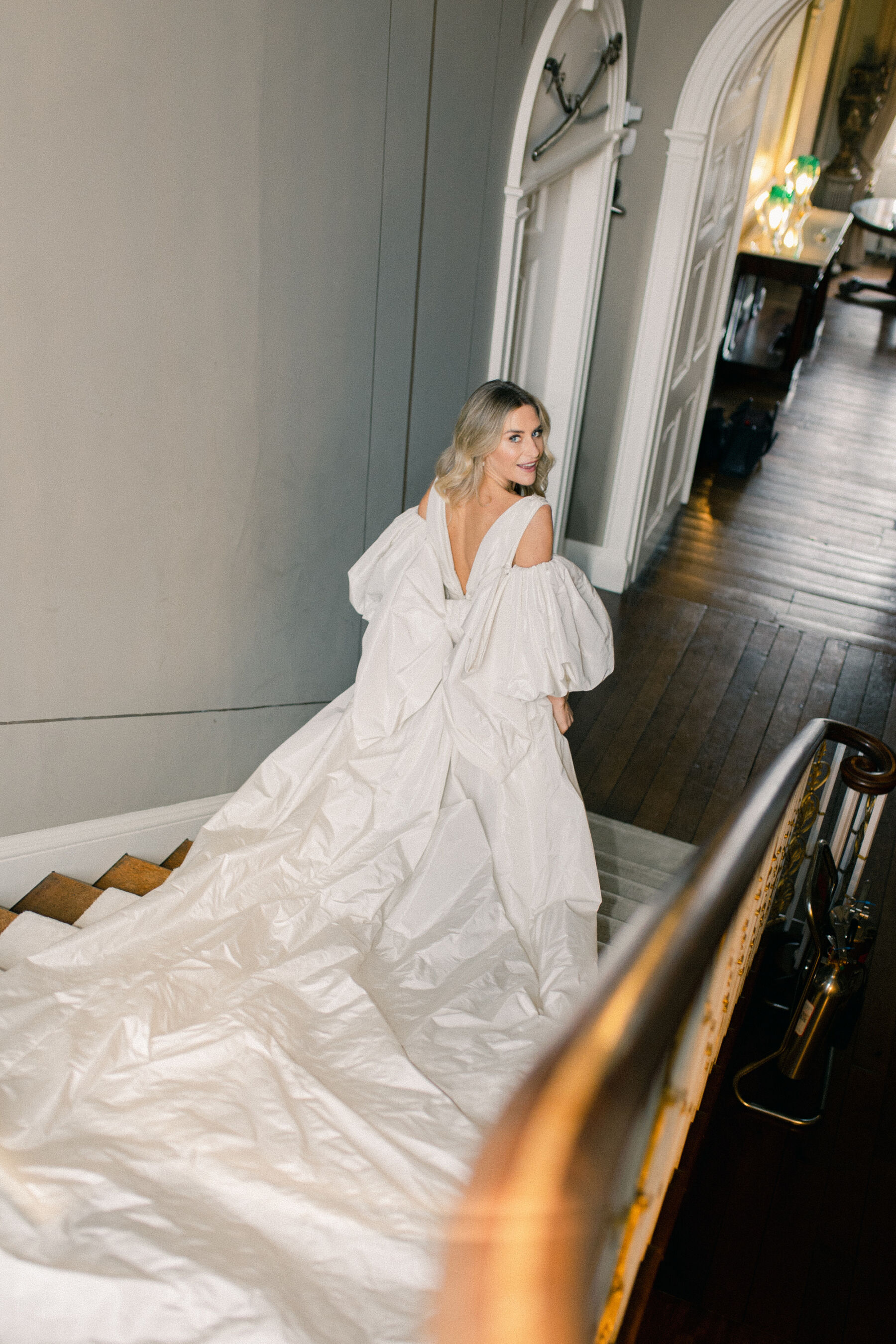 Pale Baby Blue Spring Wedding Inspiration at Avington Park, Featuring Halfpenny London 16 Bride walking down the stairs at Avington Park wedding venue in Hampshire, wearing detachable puff sleeves and huge bow - bridal gown by Halfpenny London.