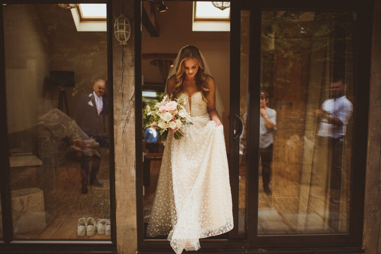Intimate photograph of a bride gently looking down to catch her step, as she gathers her Hopkins wedding dress by Pronovias to step through the doors at Tythe wedding venue, Oxfordshire. She carries a pale pink pastel colour summer wedding bouquet.. Matt Penberthy Photography.