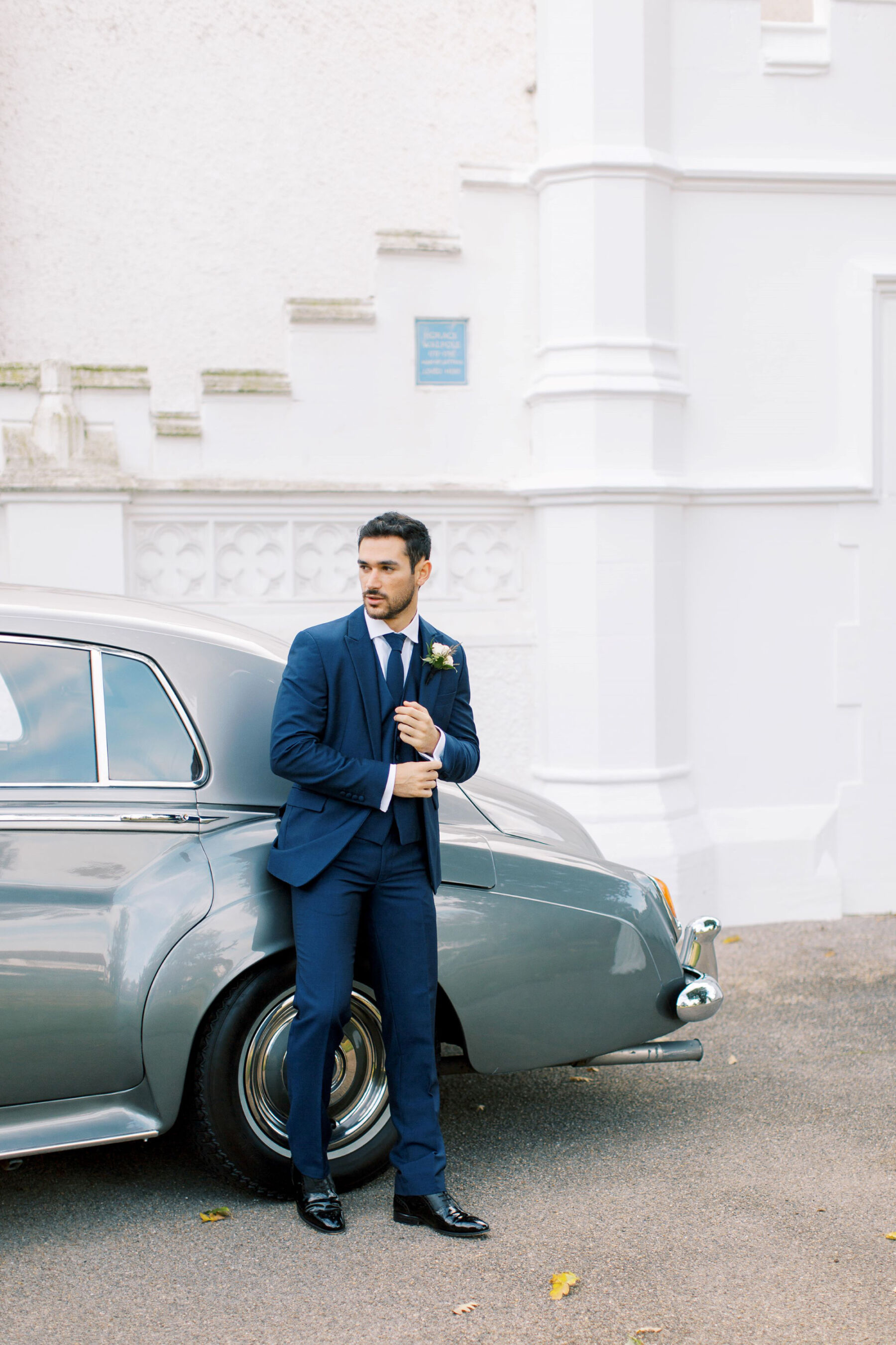 Gothic Architecture Inspired Wedding Styling, at Strawberry Hill House 21 Stylish groom in a blue suit leans against a Rolls Royce that is parked outside Strawberry Hill House & Garden. Helen Warner Photography.