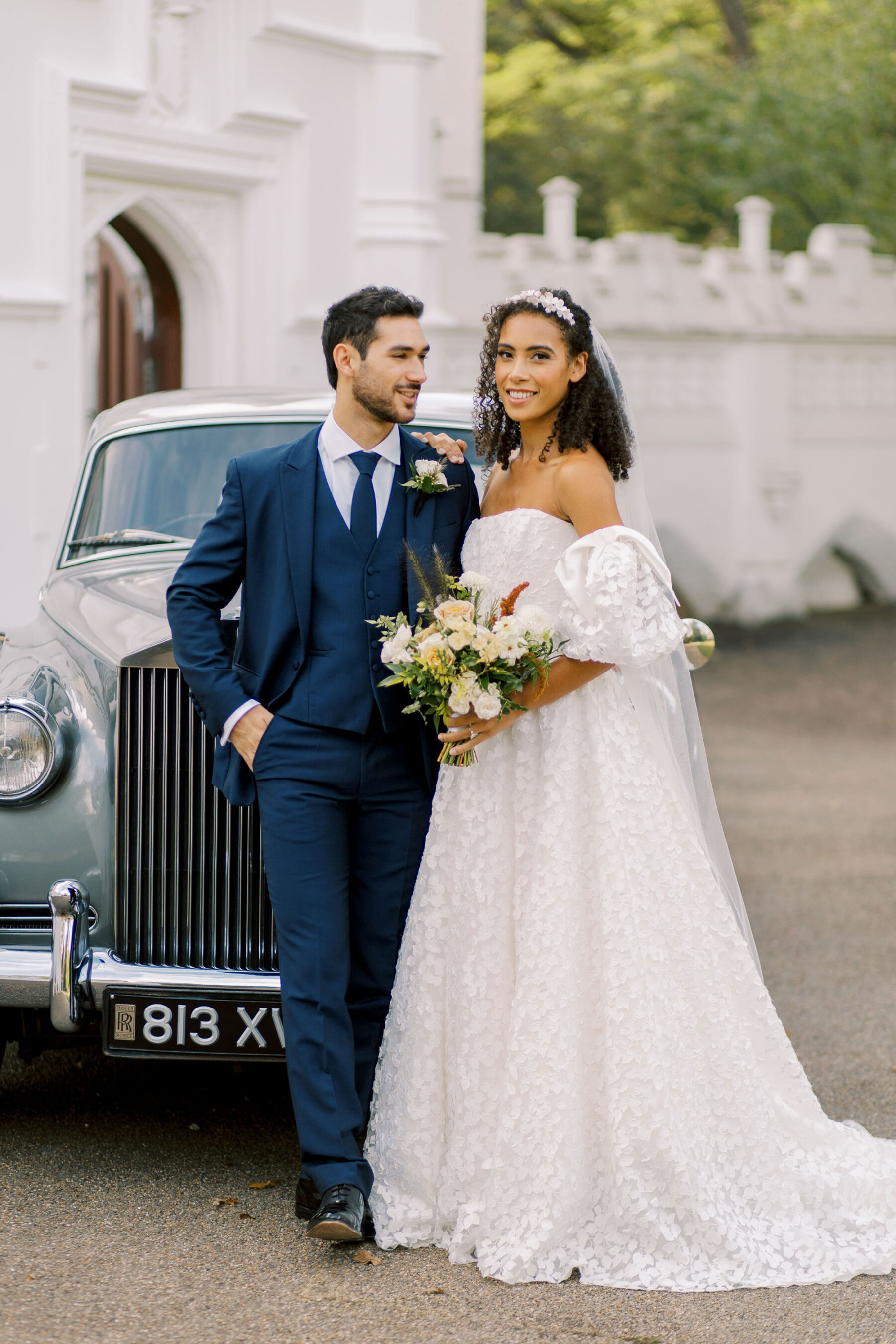Gothic Architecture Inspired Wedding Styling, at Strawberry Hill House 24 Stylish modern bride in a puff sleeve wedding dress by Sassi Holford, smiling with her groom in a suave blue suit. The groom leans against a Rolls Royce that is parked outside Strawberry Hill House & Garden. Helen Warner Photography.