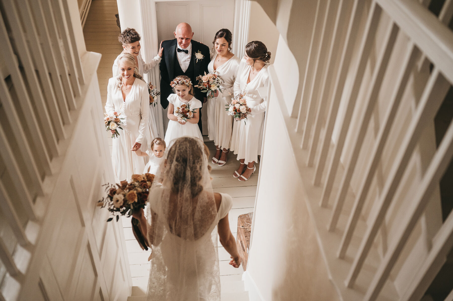 A Stylish Autumn Wedding at Lyde Court: Suzanne Neville by Day & Rime Arodaky Mini Dress Glamour By Night 9 Bride walking down the stairs and being greeted by her bridesmaids, wearing white dresses by Ghost Fashion, a flower girl in a floral crown and her father wearing a black tie tuxedo.