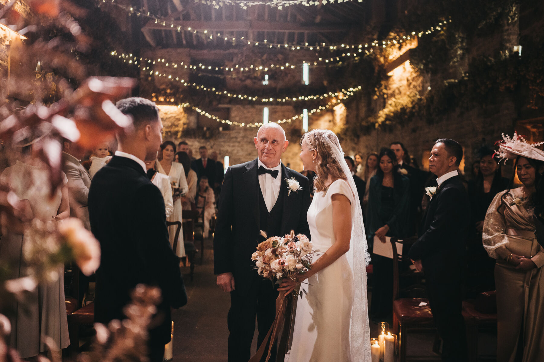 A Stylish Autumn Wedding at Lyde Court: Suzanne Neville by Day & Rime Arodaky Mini Dress Glamour By Night 15 Lyde Court wedding, bride in Suzanne Neville carrying an Autumn wedding bouquet arriving to the ceremony with her father in black tie. Twinkling fairy lights hang from the wooden beams of the barn ceiling behind them.