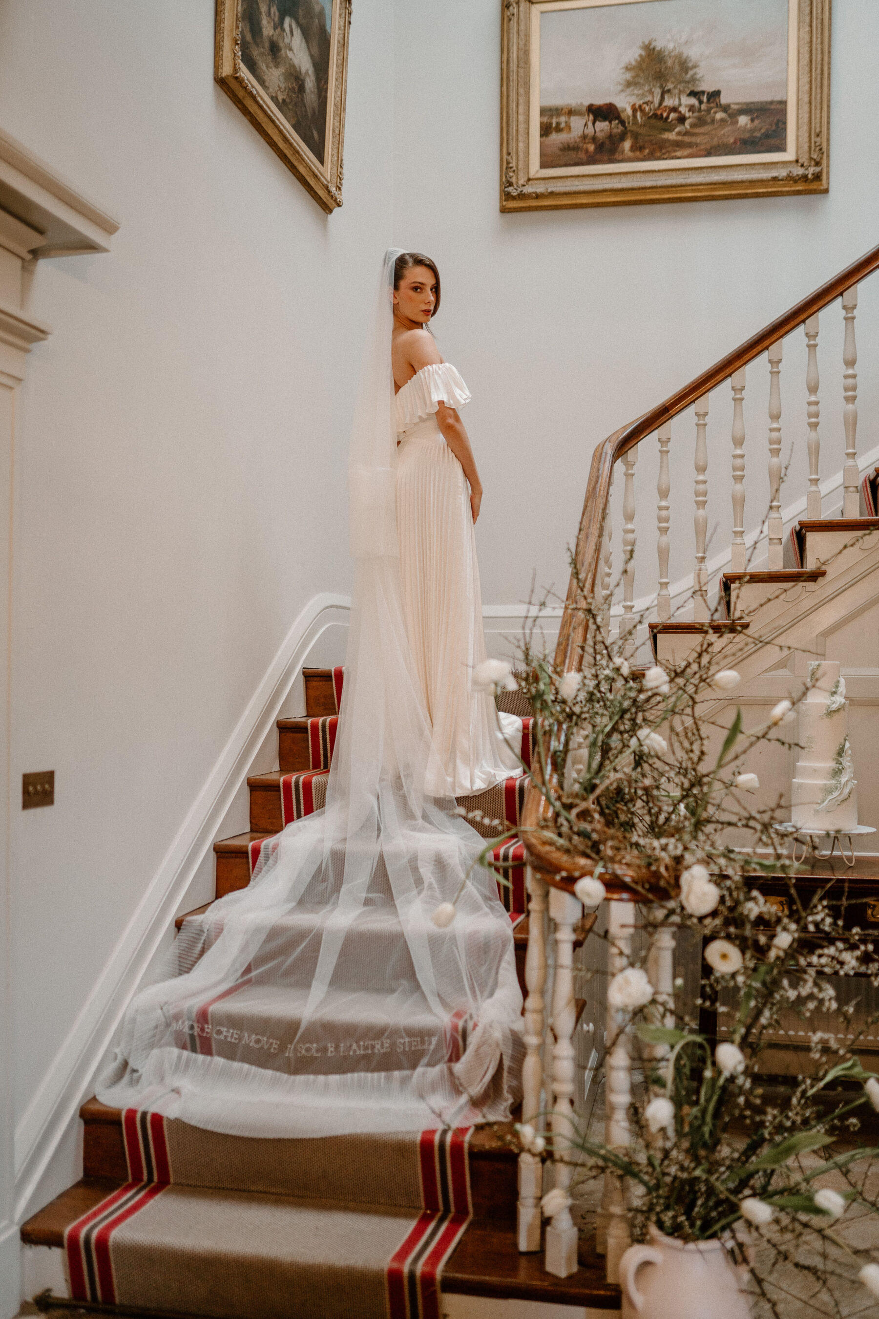 Bride in a pleated bridal two-piece standing on a staircase and looking back to the camera. She wears an embroidered Rebecca Anne Designs veil.