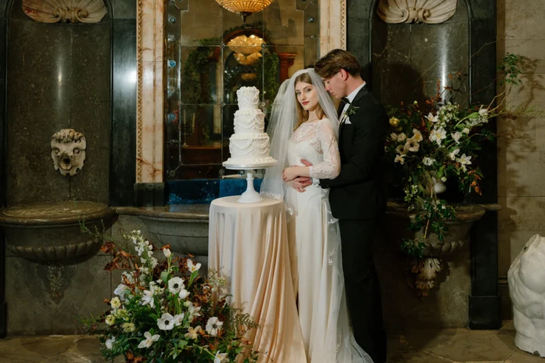 Bride in a long-sleeved lace wedding dress and veil with her groom beside a tiered white cake and spring flowers at Thornbridge Hall