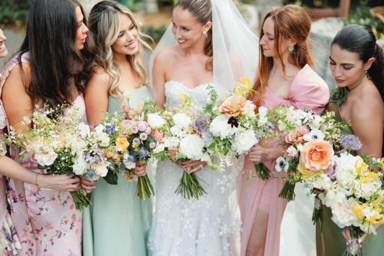 Bride with four bridesmaids holding floral bouquets at outdoor wedding ceremony.